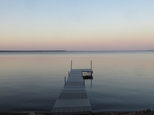 A calm lake at dusk with a wooden dock extending into the water, reflecting soft pastel colors in the sky.