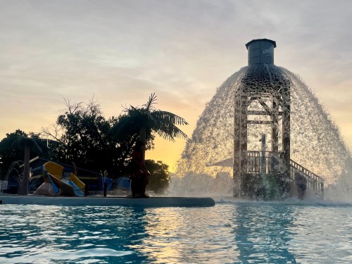 A water park scene at sunset, featuring a large water fountain and colorful slides beside a pool.