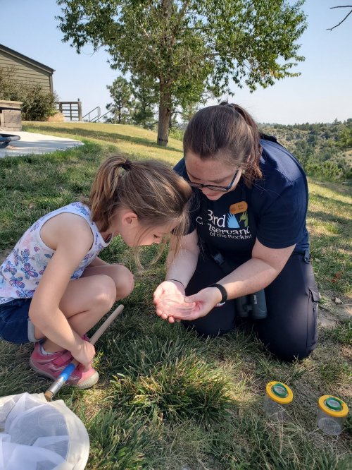 A woman and a young girl examine a small object in the girl's hands while sitting on the grass outdoors.