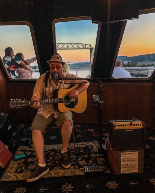 A musician plays guitar and sings inside a venue with large windows, showcasing a sunset and people enjoying the view.
