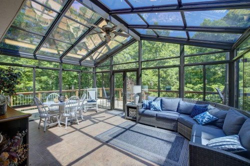 Bright sunroom with glass walls, featuring a cozy sofa, white chairs, and a view of lush greenery outside.
