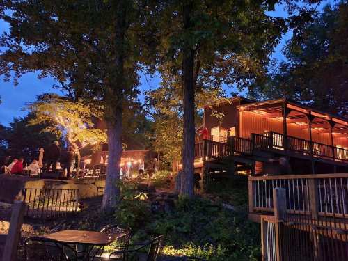A cozy outdoor restaurant scene at dusk, with warm lights, trees, and people enjoying the atmosphere.
