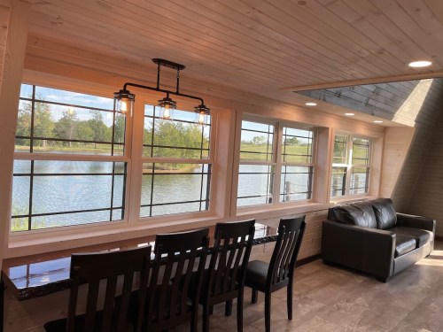 Cozy interior with a dining area by large windows overlooking a serene lake and greenery. Modern lighting and furniture.