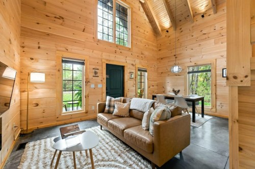 Cozy wooden interior of a cabin with a sofa, coffee table, and dining area, featuring large windows and natural light.
