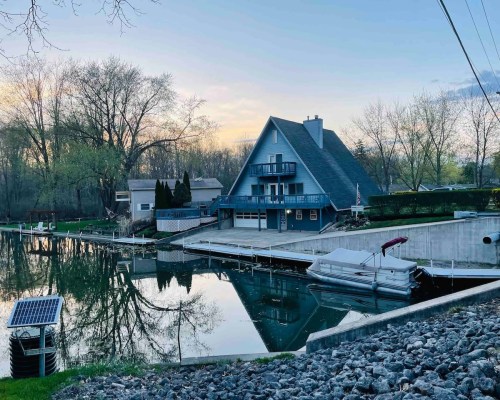 A blue A-frame house by a calm lake, surrounded by trees, with a dock and a boat moored nearby.