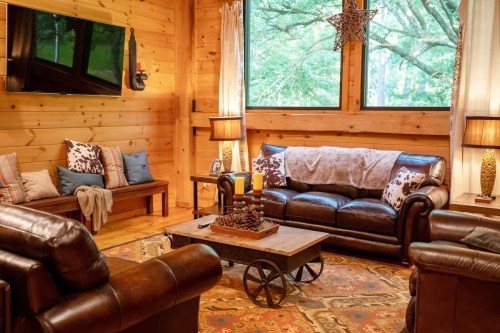 Cozy living room with leather sofas, wooden walls, a coffee table, and large windows overlooking greenery.