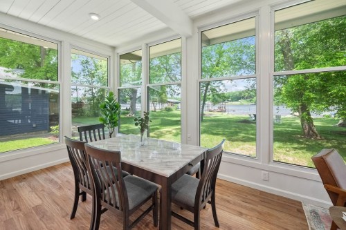Bright sunroom with a marble dining table and chairs, surrounded by large windows overlooking a green yard and lake.