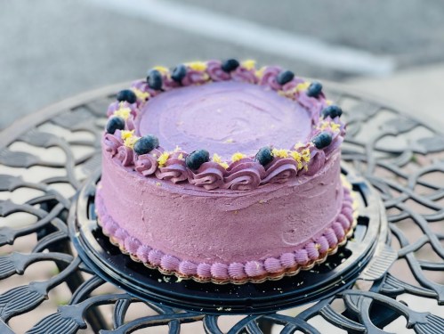 A round purple cake decorated with blueberries and yellow sprinkles, placed on an ornate black table.