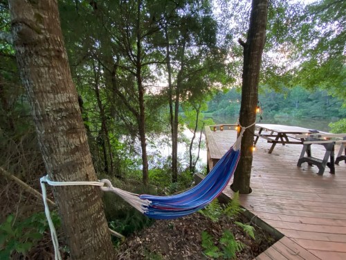 A colorful hammock hangs between trees near a wooden deck by a serene lake, surrounded by lush greenery.