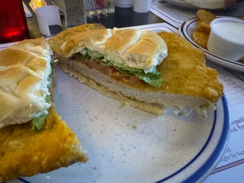 A close-up of a fried chicken sandwich with lettuce and tomato, served on a plate with a side of dipping sauce.