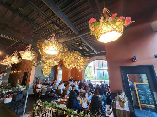 A vibrant restaurant interior with floral light fixtures, patrons dining, and a large window letting in natural light.