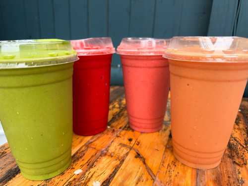 Four colorful smoothie cups—green, red, pink, and orange—sitting on a wooden table against a blue background.