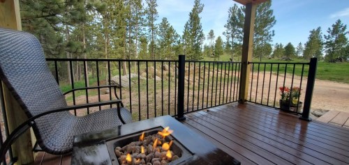A cozy outdoor deck with a fire pit, a chair, and a view of trees and a dirt path in the background.