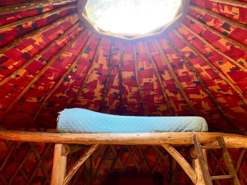 Interior of a yurt with a colorful patterned ceiling and a blue mattress on a lofted platform.