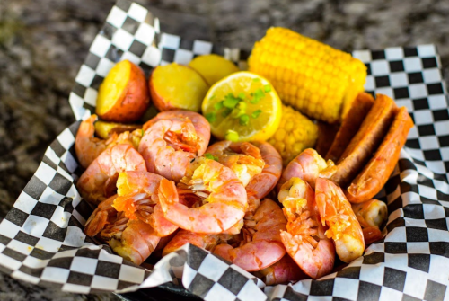 A basket of shrimp, corn, potatoes, and sausages, garnished with lemon and served on a checkered paper.