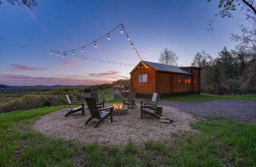 A cozy cabin surrounded by chairs and a fire pit, with string lights and a scenic sunset in the background.