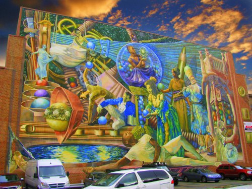 Colorful mural on a building depicting various figures and abstract elements against a dramatic sky.