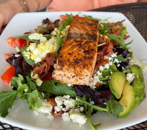 A fresh salad topped with grilled salmon, bacon, avocado, tomatoes, and crumbled cheese on a white plate.
