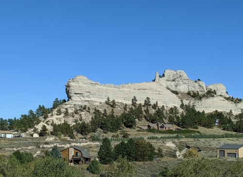 A rocky cliff rises above a green landscape, with houses nestled at its base under a clear blue sky.