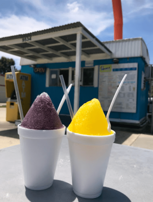 Two cups of colorful snow cones, one purple and one yellow, sit on a table in front of a blue building with an open sign.