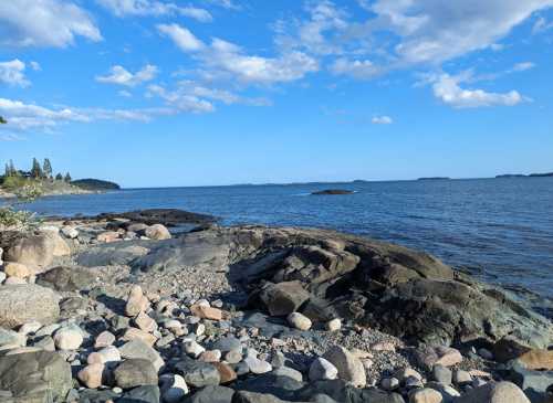 A rocky shoreline with smooth stones, calm blue water, and a clear sky dotted with clouds.