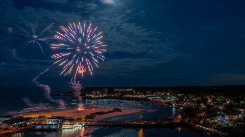 Colorful fireworks illuminate the night sky over a coastal town, reflecting on the water below.