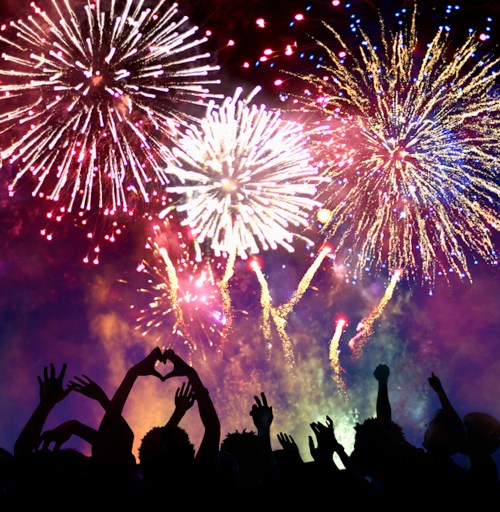Silhouetted crowd with raised hands against a vibrant fireworks display in the night sky.