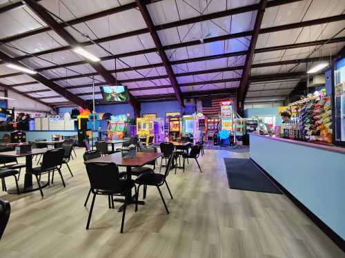 Brightly lit arcade with games, seating area, and a snack bar, featuring colorful machines and a spacious layout.