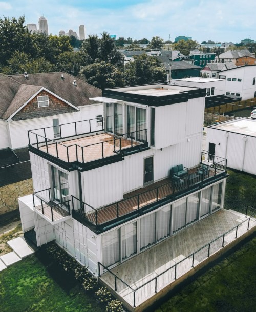 A modern, multi-level house made from shipping containers, featuring a rooftop deck and large glass windows.