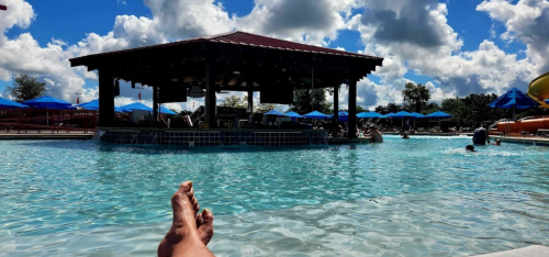 A sunny pool scene with a person’s feet in the foreground, a shaded cabana, and blue umbrellas in the background.