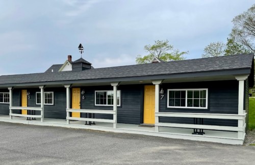 A row of dark blue motel rooms with yellow doors, set against a cloudy sky and green trees in the background.
