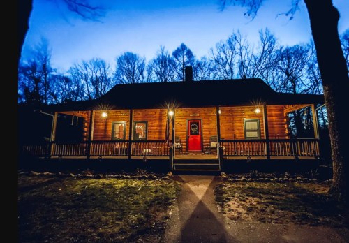 A cozy wooden cabin at dusk, illuminated by porch lights, surrounded by bare trees.
