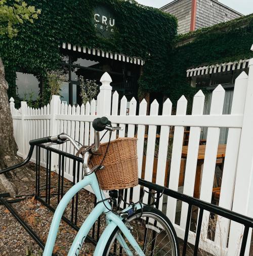 A light blue bicycle with a wicker basket parked by a white picket fence in front of a cafe covered in greenery.