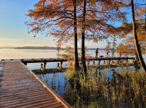 A serene lake scene with autumn trees, a wooden dock, and a person walking along the water's edge.