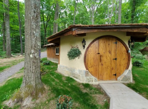 A charming round cottage with wooden doors, nestled in a lush green forest. Pathway leads to the entrance.