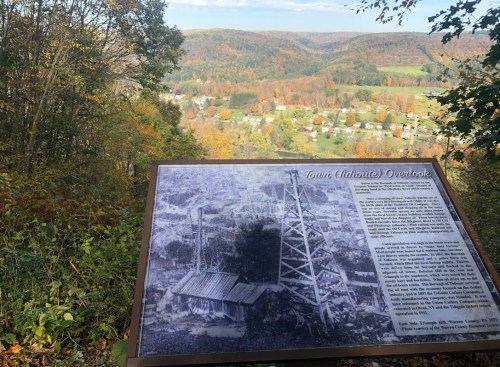 A scenic overlook with a historical sign, showcasing a valley surrounded by autumn foliage and a town below.
