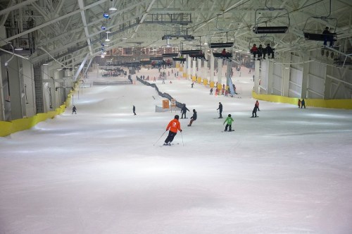 Indoor ski slope with skiers and snowboarders on a snowy surface, surrounded by a spacious, well-lit facility.