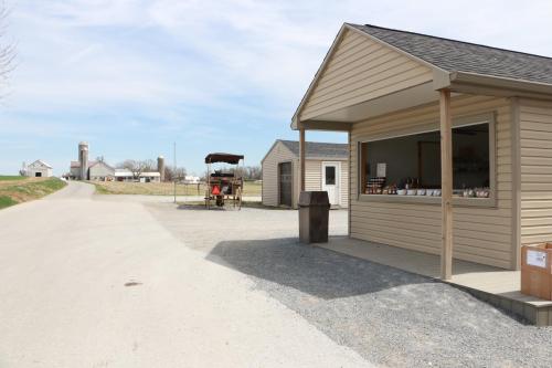 A small building with a porch on a gravel road, with farm buildings and a wagon in the background under a blue sky.