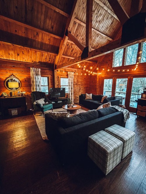 Cozy wooden living room with string lights, large windows, and comfortable seating, overlooking a snowy landscape.