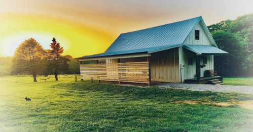 A charming house with a metal roof sits on a grassy field at sunset, surrounded by trees and a peaceful landscape.