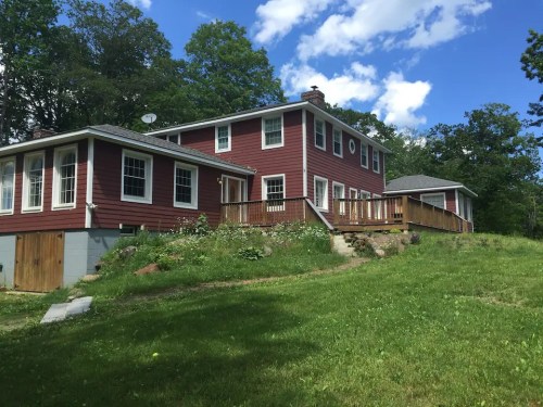 A red two-story house with a wooden deck, surrounded by green grass and trees under a partly cloudy sky.