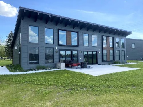 Modern two-story building with large windows, surrounded by green grass and a patio area. Bright blue sky in the background.
