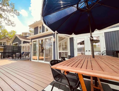A spacious deck with a wooden table, black chairs, and a large umbrella, overlooking a modern house and trees.