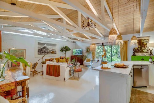 Bright, airy living space with wooden beams, cozy seating, and natural decor, featuring large windows and greenery.