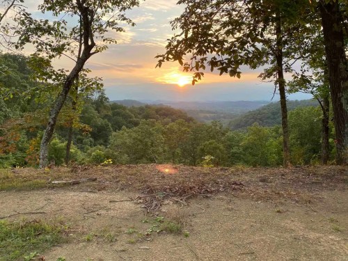 A serene sunset over rolling hills, framed by trees, with a warm glow illuminating the landscape.
