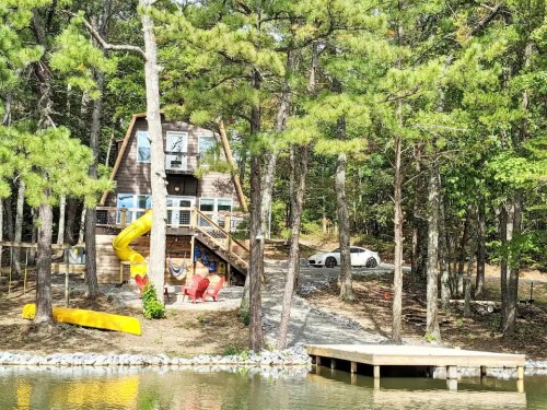A wooden cabin by a lake, surrounded by trees, with a yellow slide, red chairs, and a dock.