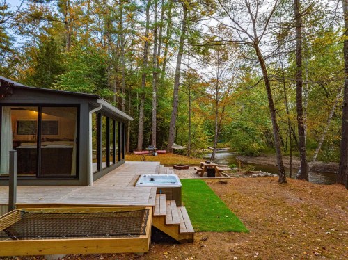 A cozy cabin by a river, featuring a hot tub and surrounded by autumn foliage and trees.