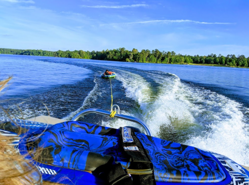 A person is being towed on a tube behind a boat on a calm lake, surrounded by trees and a clear blue sky.