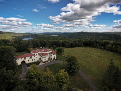 Aerial view of a large mansion surrounded by lush greenery and rolling hills under a partly cloudy sky.