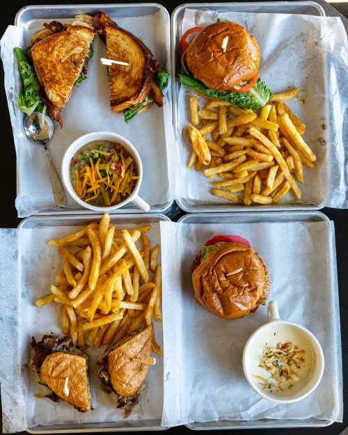 A spread of two sandwiches, two burgers, fries, and a salad in metal trays on a table.